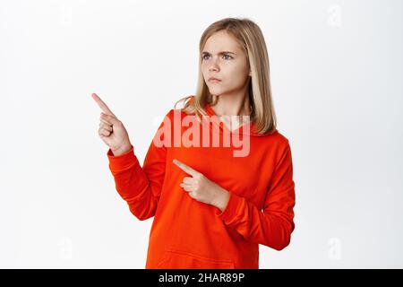 Sulking little girl staring at upper left corner, pointing at logo advertisement with doubtful, skeptical face expression, standing over white Stock Photo