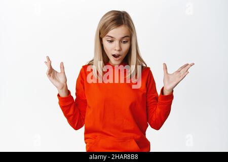 Surprised and shocked little blond girl looks down startled, seeing something lying below, drop on floor, standing over white background Stock Photo
