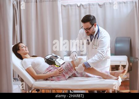 Young Caucasian doctor dressed in white uniform putting gauze on patient's knee injury. Stock Photo