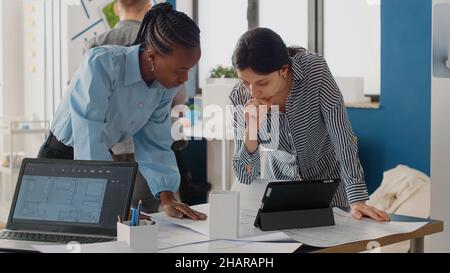 Close up of diverse women analyzing blueprints plan with digital tablet, doing teamwork for project. Architects working on construction layout and structure for urban development. Stock Photo