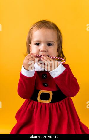 Portraite Cute Cheerful Chubby Baby Girl in Santa Suite Holding Eating Caramel Candy At Yellow Background. Child Play Christmas Scene Celebrating Stock Photo