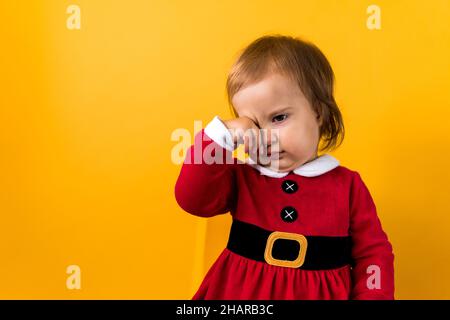 Banner Portraite Cute Happy Cheerful Chubby Baby Girl in Santa Suit Sleepy Rubs Eyes At Yellow Background. Child Play Christmas Scene Celebrating Stock Photo