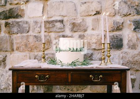 Bunk wedding cake decorated with green branches stands on a wooden table near a stone wall Stock Photo