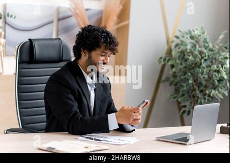 Satisfied successful young adult Indian, top manager, business man sitting at table, using smartphone while working, browsing Internet or news, answering e-mail, texting with friends, smiling Stock Photo