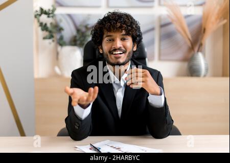 Smart attractive successful Indian business leader, mentor or manager talking to an employee or client during video communication, brainstorming, gesturing with his hands, look at camera, smile Stock Photo