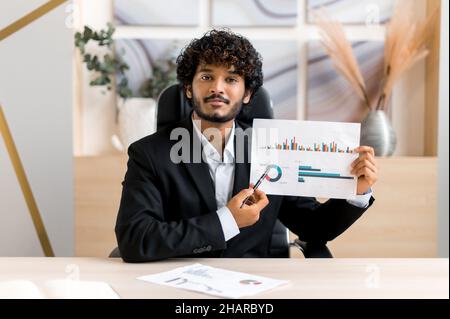 Smart successful curly-haired Indian business leader, financial mentor, corporate manager talk to colleague or client on conference call, virtual meeting, brainstorming, holds documents,look at camera Stock Photo