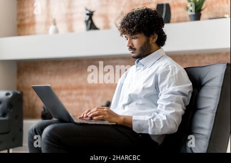 Concentrated serious curly-haired indian curly haired man, sitting in armchair, using laptop, texting with friends, colleagues or family, chatting on social media, answering e-mail Stock Photo