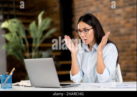 Caucasian businesswoman suffered financial losses. Upset shocked young adult brunette broker with glasses sitting in office with laptop surprised by news or message, collapse, gesturing with hands Stock Photo