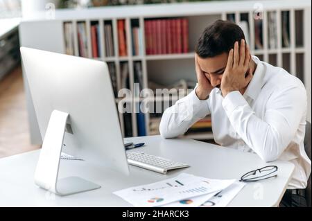 Overwhelmed exhausted Indian company owner, manager or office worker, sitting at his desk with closed eyes, rubbing his head, feeling stressed and tired from overtime, needs rest Stock Photo