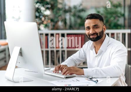 Friendly smart confident businessman of Indian ethnic, manager or ceo with beard sitting in office, wearing white shirt, using computer, texting with clients, taking notes, looking at camera, smiling Stock Photo