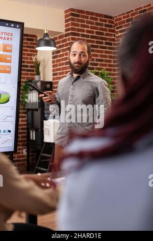 Entrepreneur man pointing at monitor explaining management statistics to businessteam working at company presentation in startup office. Diverse businesspeople planning business presentation Stock Photo