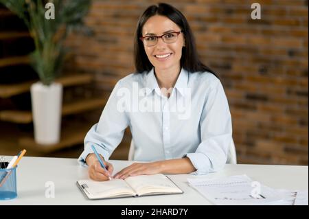 Friendly confident pretty smart young adult brunette Caucasian woman, top manager, broker, sitting at the table in the office, talking with colleagues, online partners, taking notes, smiling Stock Photo