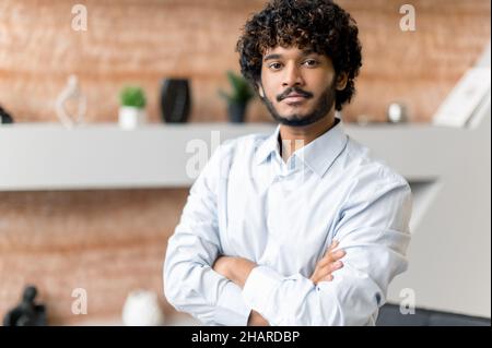 Portrait attractive successful modern curly haired Indian or Arabian man with beard, office worker, freelancer, top manager, wearing shirt, standing in office with his arms crossed, looking at camera Stock Photo