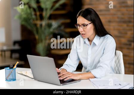 Serious puzzled young adult caucasian brunette business woman, ceo or top manager wearing glasses sitting in office, using laptop, analyzing financial market, preparing business strategy or report Stock Photo