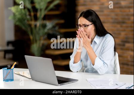 Upset shocked young adult caucasian brunette business woman, CEO or broker wearing glasses sitting in office at laptop, surprised by news or message, financial loss, collapse. Emotional stress Stock Photo