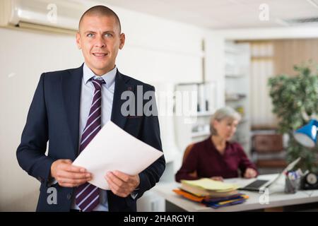 Confident smiling entrepreneur standing in office with paper documents Stock Photo
