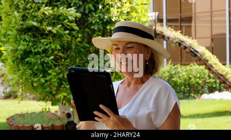 managing Senior female blonde businesswoman in hat uses tablet pc, digital tablet for business work or study in her own green garden. woman aged 50-55 Stock Photo