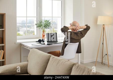 Relaxed man rests, stretches and puts his hands behind his head while sitting at home workplace. Stock Photo