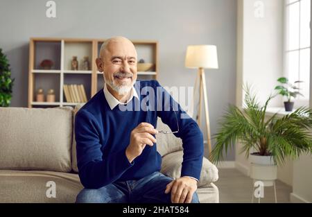 Happy, cheerful senior man sitting on sofa at home, holding his glasses and smiling Stock Photo