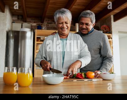 Multi-cultural elderly couple laughing in modern kitchen. Healthy retired lifestyle at home, preparing fruit salad.  Stock Photo