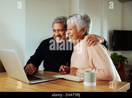 Happy multi-cultural elderly couple smiling at family over online call. Sitting in at modern kitchen counter at home. Stock Photo