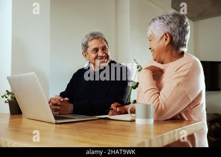 Multi-cultural elderly couple smiling at each other. Sitting at modern kitchen counter with laptop. Stock Photo