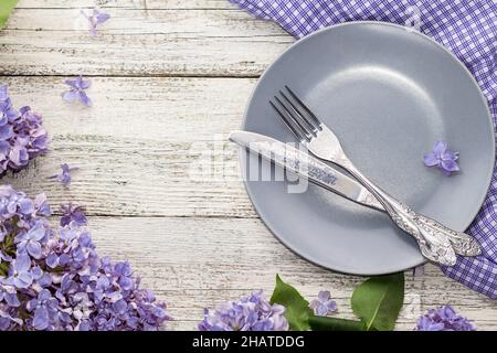 spring table setting with plate fork and knife decorated with lilac flowers on white wooden background. Top view with copy space in very peri trendy c Stock Photo