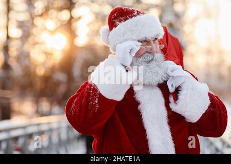 Horizontal medium close-up portrait of Santa Claus wearing eyeglasses walking outdoors carrying sack with gifts looking away