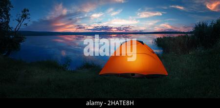 Internally lit orange tent on shore of lake under dramatic sunset sky