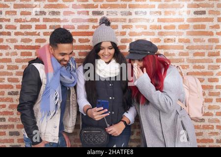 Smiling young woman showing video on cellphone to multiethnic friends while stylish female with red hair pointing at screen against brick wall Stock Photo