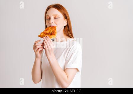 Studio portrait of cheerful young woman with closed eyes appetite eating delicious pizza standing on white isolated background. Stock Photo