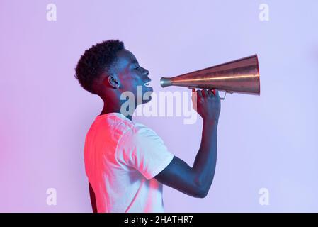 Side view of cheerful young black male millennial in t shirt smiling while talking with metal vintage megaphone against purple background Stock Photo