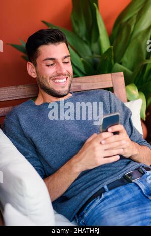 Positive bearded male text messaging on cellphone while sitting in wooden armchair with pillow in room with potted green plant Stock Photo
