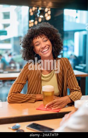 Cheerful African American female with short curly hair in casual clothes sitting at wooden bar counter with glass of beer and laughing in pub in dayti Stock Photo