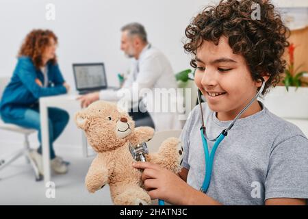 Happy male child listening his plush bear toy using stethoscope standing in doctor's office with his mother during pediatrician consultation Stock Photo