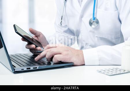Male doctor with laptop and cellphone at desk in medical office. Physician using laptop at desk in clinic working on computer at room office. Doctor u Stock Photo