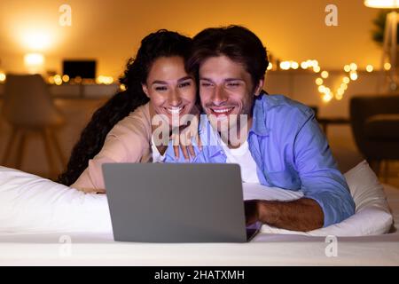 Smiling Couple Using Laptop Watching Funny Film In Bedroom Stock Photo