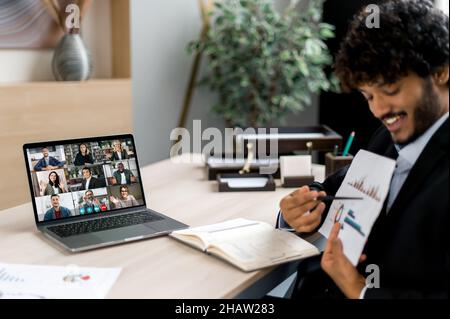 Positive successful Indian businessman, top manager or coach, uses laptop for online conference with employees, holds briefing, shows documents and charts, on screen are multiracial business people Stock Photo