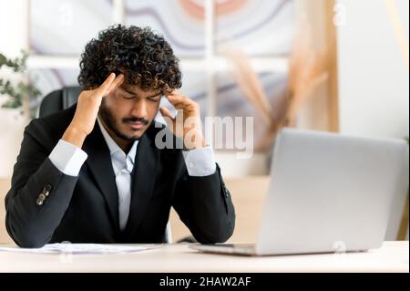 Exhausted tired young adult curly-haired Indian businessman, broker or manager sits at the table in the office, massaging temples, he needs rest, is stressed, headache, migraine. Feelings unwell Stock Photo