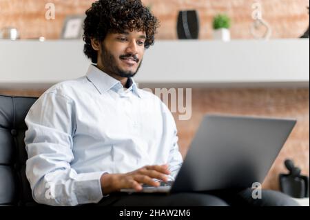 Smart focused confident Indian freelancer, top manager or stock broker, in stylish clothes with beard, uses laptop, reads news, analyzing stock market, planning, prepares financial strategy Stock Photo
