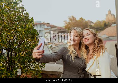 Close-up portrait of two female friends in strict suits laughing taking selfie on the terrace outside at summer street cafe on background buildings of Old Tbilisi city, Georgia. High quality photo Stock Photo