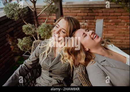 Close-up portrait of two female friends in strict gray suits laughing while sitting on wicker chairs on the terrace outside at summer street cafe. High quality photo Stock Photo