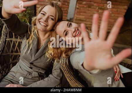 Close-up portrait of two female friends in strict gray suits laughing and closing faces with hands from the camera while sitting on wicker chairs on the terrace outside. High quality photo Stock Photo