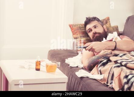 Bearded ill man lying in bed at home. Sick man lying on sofa with TV remote under a blanket at home in the living room. Stock Photo