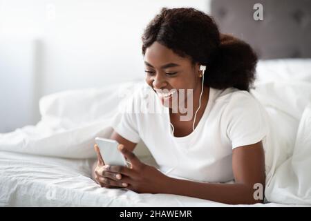 Happy young black woman laying in bed with smartphone Stock Photo