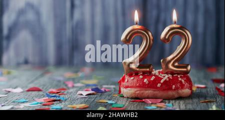 lit golden number-shaped candles forming the number 22 topping a small cake with a red frosting, on a gray rustic wooden table with some confetti, in Stock Photo