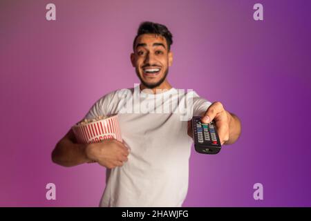 Cheerful millennial Arab guy watching TV and eating popcorn, switching channels with remote control in neon light Stock Photo