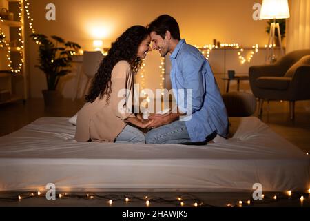 Loving Couple Holding Hands Touching Foreheads Sitting In Bedroom Stock Photo