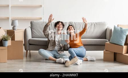 Millennial Asian couple sitting on floor among carton boxes, using digital tablet, dreaming about design of new home Stock Photo