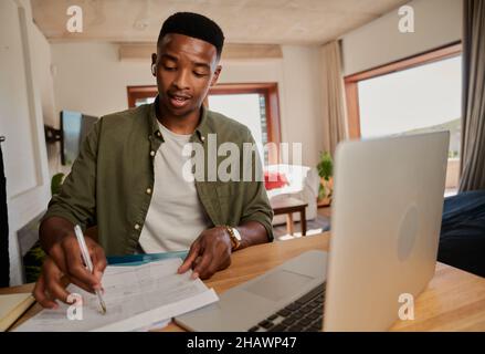 Young adult black male wearing earphones making notes while on online meeting. Sitting at kitchen counter using laptop. Stock Photo